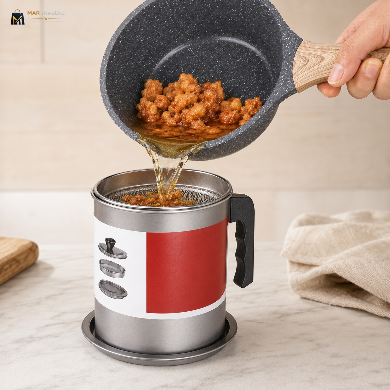 A hand holds a dark gray speckled frying pan above a stainless steel and red-and-white insulated mug with a strainer, pouring hot oil over food. The scene is set on a light marble countertop with a beige towel in the background.