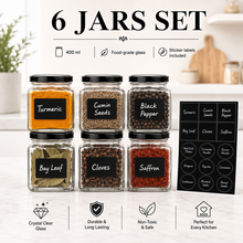 Six clear glass spice jars with black lids arranged in two rows on a light countertop, labeled with spices including turmeric, cumin seeds, black pepper, bay leaf, cloves, and saffron. A black sticker label sheet is shown beside the jars, with text at the top reading '6 JARS SET' and feature icons below.