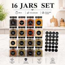 A 16-jar spice and seasoning set displayed in a bright kitchen setting. The set includes 16 clear glass jars with black lids, filled with various spices and seeds, and a sheet of 16 matching black circular labels with white text.