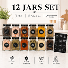 A 12-jar spice set arranged in two rows on a light countertop, with a blurred kitchen background. The jars are clear food-grade glass with black lids and labeled with black circular stickers showing spice names such as black pepper, pink salt, red chili flakes, cumin seeds, turmeric powder, coriander seeds, masala salt, oregano, paprika, bay leaves, garlic powder, and cloves.