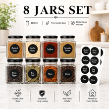 An 8-piece glass spice jar set arranged in two rows on a kitchen counter, with black lids and round black labels. The jars contain mustard seeds, garlic flakes, saffron, coriander powder, black pepper, bay leaf, mace, and cloves.