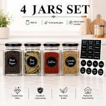Four clear glass spice jars with black lids and black label stickers, shown on a light marble surface with a softly blurred kitchen background. The jars are labeled Black Pepper, Bay Leaf, Saffron, and Coriander Seeds, with additional black seasoning labels displayed to the right.