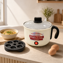 A countertop electric cooking pot with a glass lid and black handle sits on a beige kitchen counter beside a bowl of pasta, two eggs, and a black mold insert. The pot has a cream-colored body with a red power button and Chinese/English branding on the front.