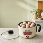 A small electric steamer pot with a handle and glass lid sits on a kitchen counter, containing several eggs. The pot is cream-colored with a black handle and base, and a red power button is illuminated.
