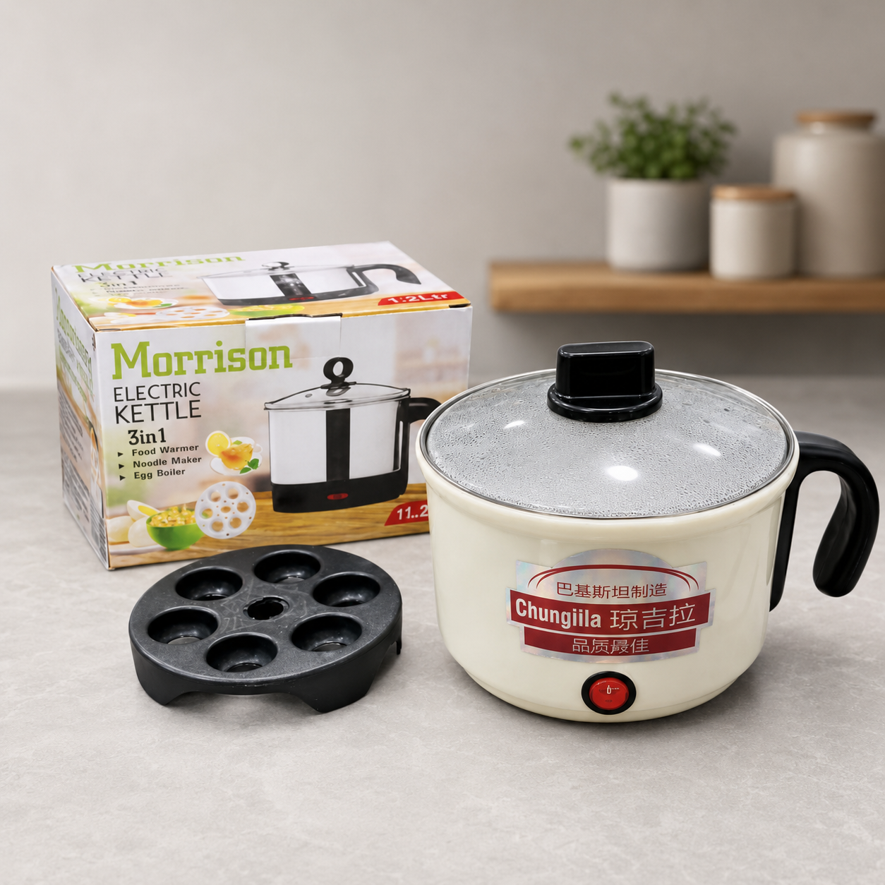 Morrison 2-in-1 Electric Kettle 5-in-1 multi-functional cooker shown with its packaging box, black non-stick mold tray, and cream-colored body with a clear glass lid on a light gray countertop.