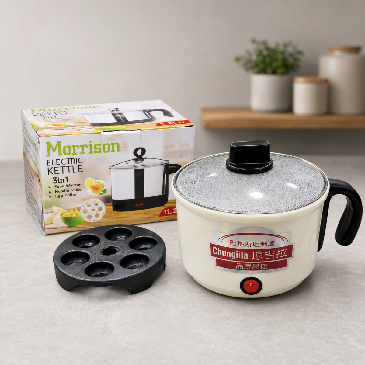 Morrison 2-in-1 Electric Kettle 5-in-1 multi-functional cooker shown with its packaging box, black non-stick mold tray, and cream-colored body with a clear glass lid on a light gray countertop.