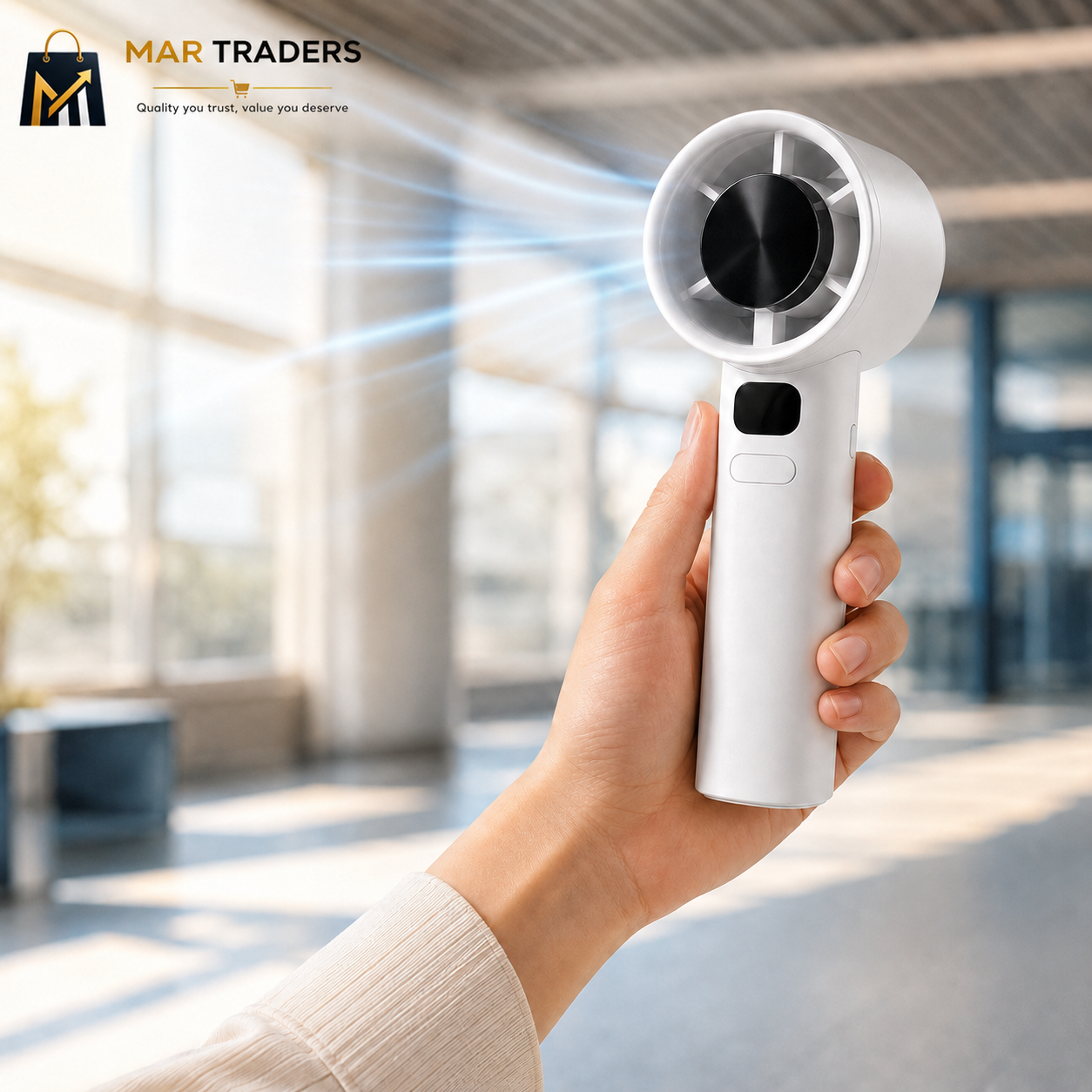 A hand holding a compact white handheld fan with a circular head, shown in a bright modern indoor setting with blurred windows and light rays. The MAR TRADERS logo appears in the top-left corner.