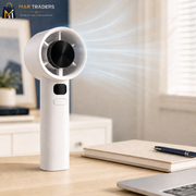 A white handheld desk fan with a black center grille, shown standing on a light wood desk in a bright home office setting. White airflow lines are illustrated coming from the fan, with a notebook, pen, and laptop visible in the background.