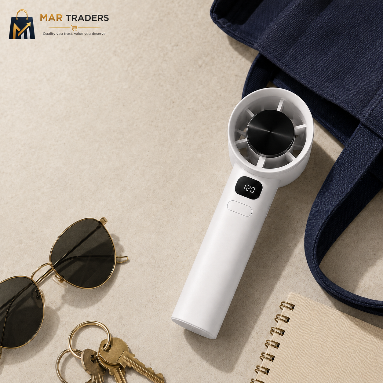 A white handheld personal fan with a digital display showing 120, styled in a flat-lay composition on a beige textured surface alongside sunglasses, keys, a notebook, and a navy blue tote bag. The MAR TRADERS logo is visible in the top-left corner.