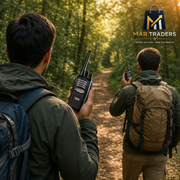 Two men hiking on a forest path, each holding a black walkie-talkie radio. One man wears a dark green jacket and blue backpack, while the other wears an olive green jacket and tan backpack.