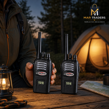 Two black Mar Traders walkie-talkies held by a person at a campsite table, with a glowing tent, lantern, and trees in the background. The Mar Traders logo is visible in the top right corner.